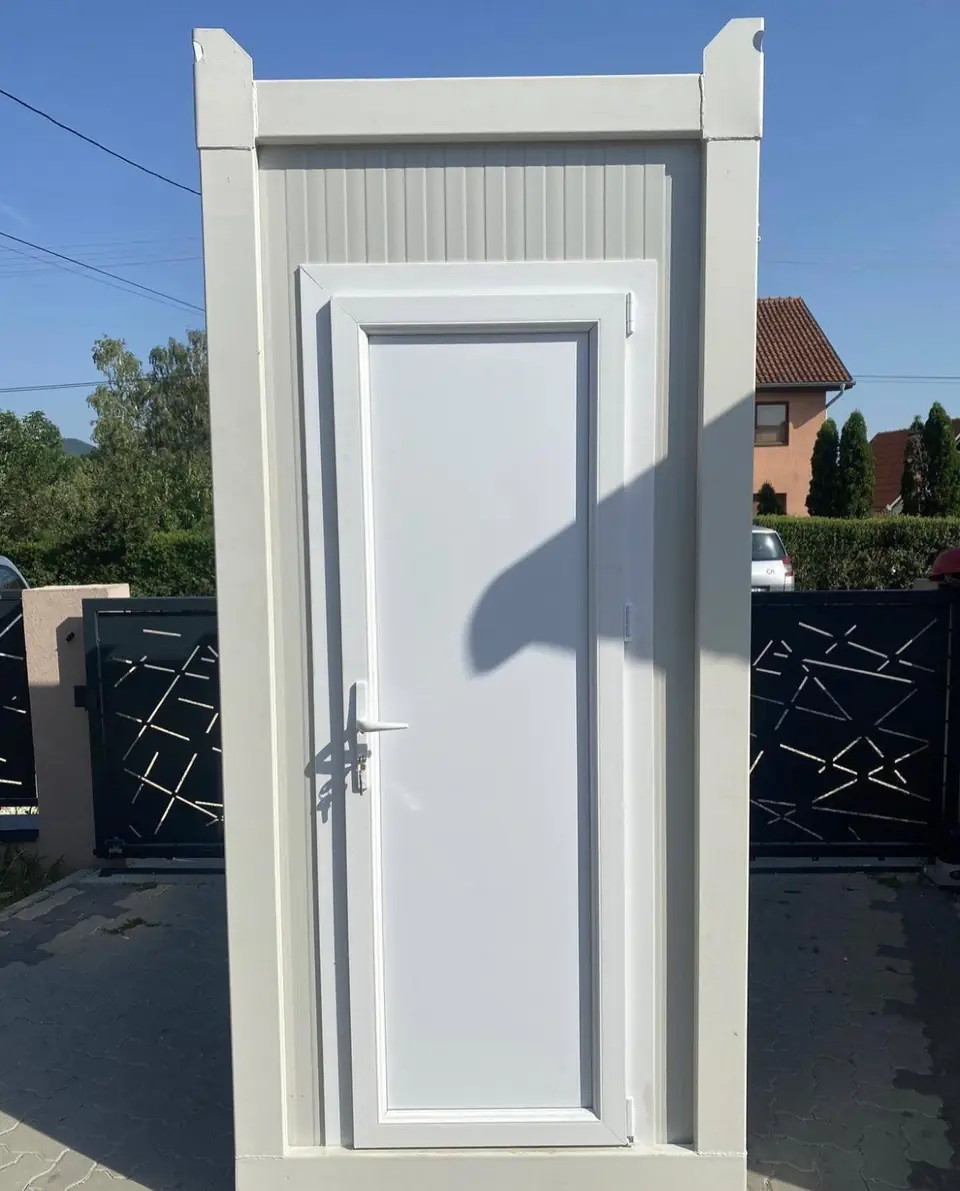 White single-stall deluxe restroom unit standing in front of a dark metal driveway gate
