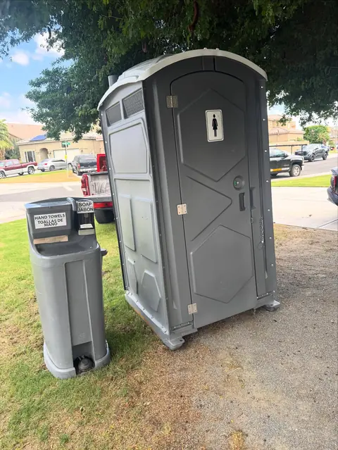 Gray porta potty with a free-standing foot-pump hand wash station on a lawn
