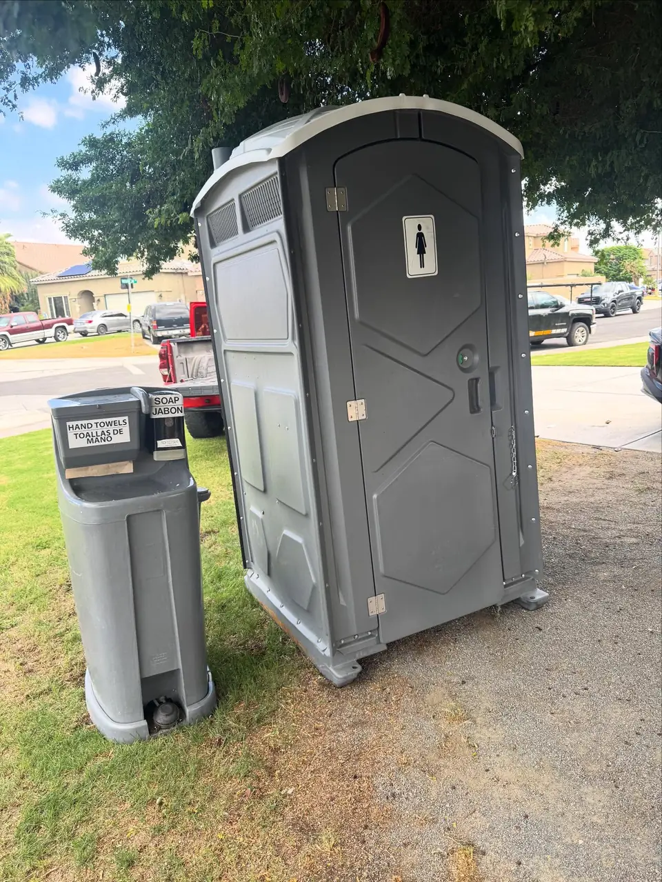 Gray porta potty beside a free-standing foot-pump hand wash station on grass