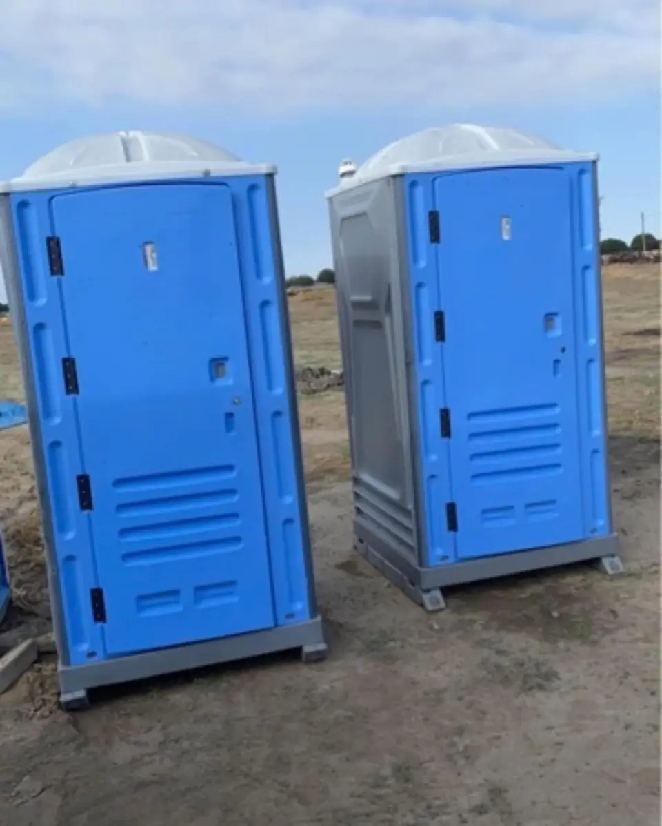 Two blue standard porta potties set on a graded job site under a clear sky